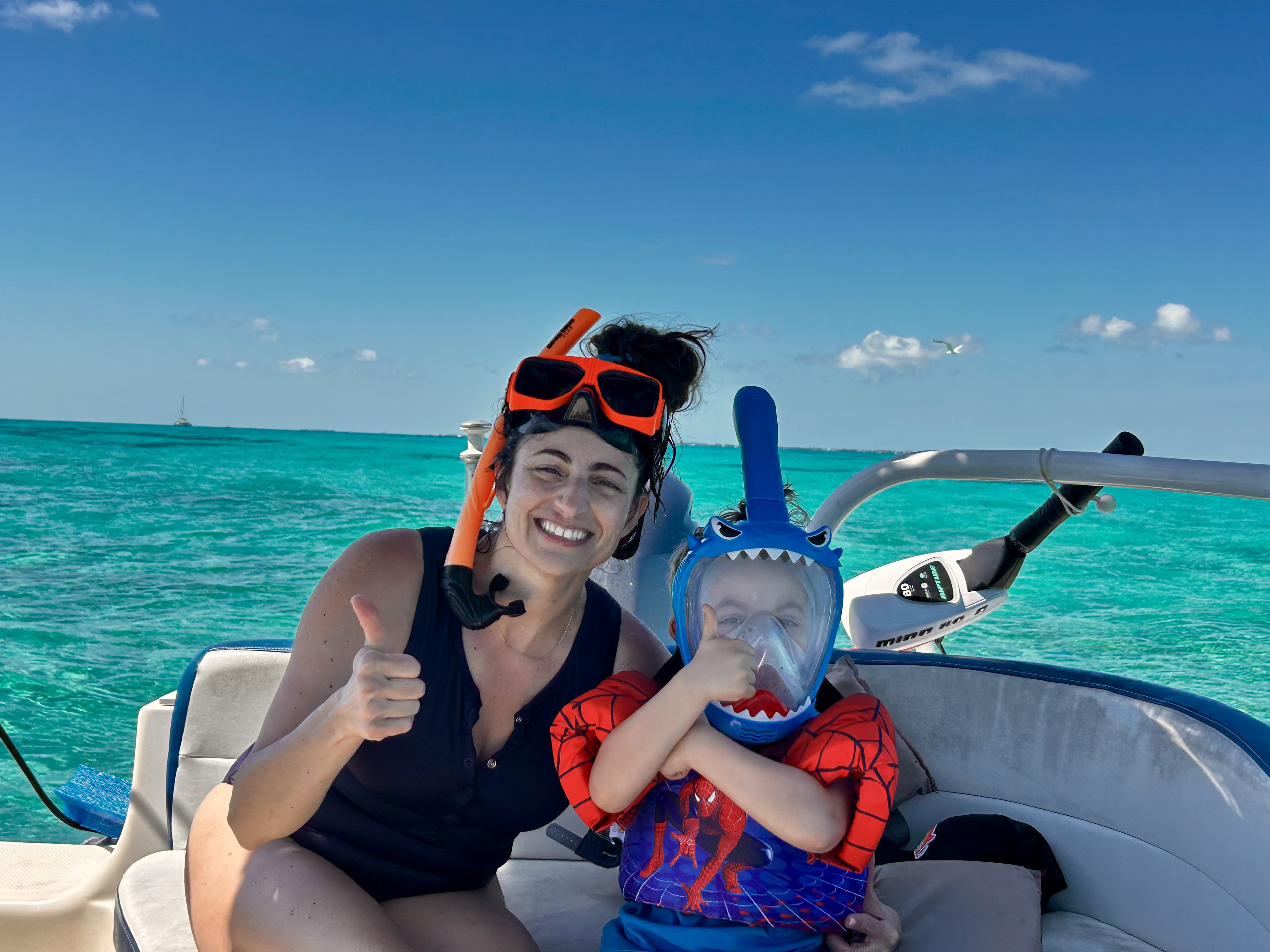 Smiling woman with orange snorkel mask resting on her forehead and a child in a blue shark full-face mask giving thumbs-up gestures while seated on a boat in sunny, clear tropical waters.